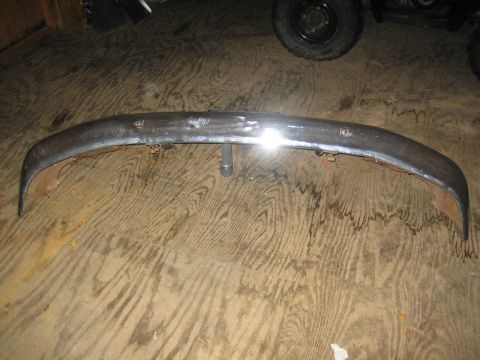 1994 Ford Bronco - A chrome bumper, likely for an older vehicle, rests on a wooden floor. It shows signs of wear, including rust and dents.
