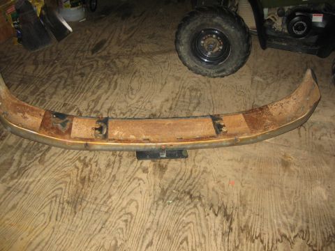 1994 Ford Bronco - A heavily rusted bumper sits on a wooden floor, with a spare tire and part of an ATV visible in the background.