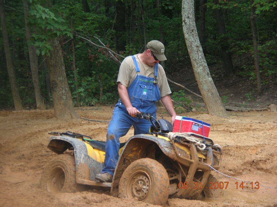 Yellow utility ATV stuck in orange mud on a wooded trail with rider working it free
