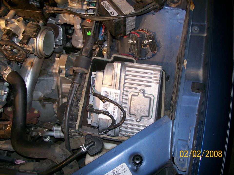 Engine bay with finned aluminum PCM module and wiring harnesses near blue inner fender
