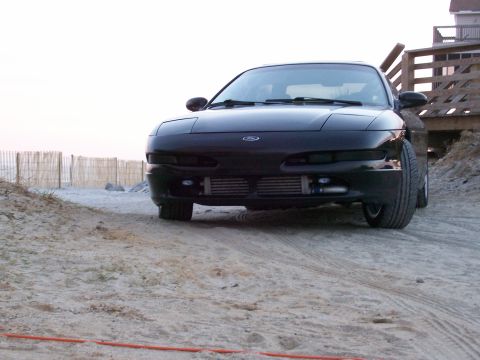 1995 Ford Probe - A black Ford Probe is parked on a sandy area near a beach with a wooden structure in the background.