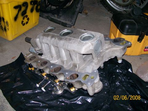 1995 Mercury Grand Marquis - A cast-aluminum V8 intake manifold sits upside-down on black plastic sheeting on a garage floor, with the fuel rail and injectors still ins…