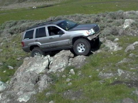 1999 Jeep Grand Cherokee - A silver Jeep Grand Cherokee is shown driving over large rocks on a grassy, hilly landscape.