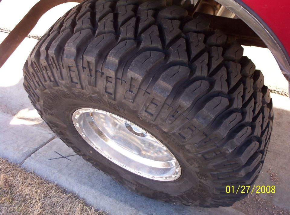 Close-up of a large, knobby off-road tire on a shiny rim. Some red vehicle body is visible above
