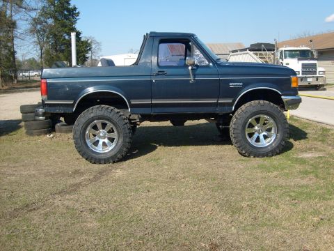 1989 Ford Bronco - This lifted Ford Bronco is shown from the side with large off-road tires and a clear blue sky overhead.