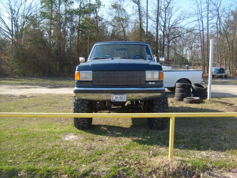 1989 Ford Bronco - A lifted dark blue pickup truck is parked outdoors with a stack of tires nearby and another vehicle in the background.