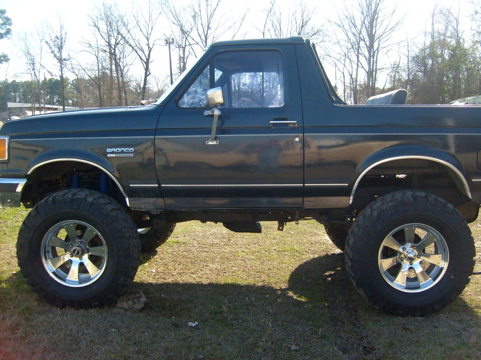 Lifted 1989 Ford Bronco, side view. Dark blue paint, chrome wheels, off-road tires