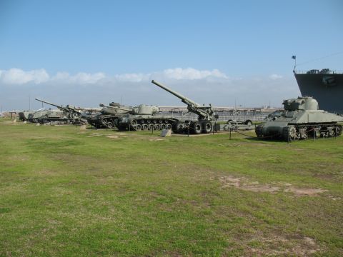 1997 Ford Crown Victoria - A collection of military vehicles, including tanks and artillery, are displayed on a grassy field with a large ship visible in the distance.