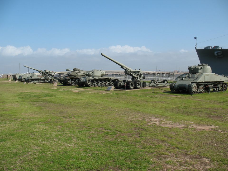 Row of tanks and artillery pieces with a large ship in the background