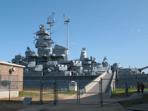 1997 Ford Crown Victoria - A large battleship is docked, with a ramp providing access to its upper levels. A chain-link fence surrounds the area.