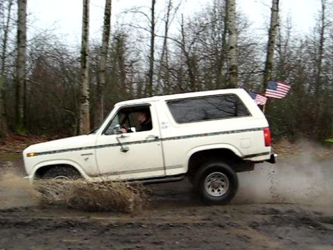 1980 Ford Bronco - White 1980 Ford Bronco kicks up mud while driving off-road with American flags attached.