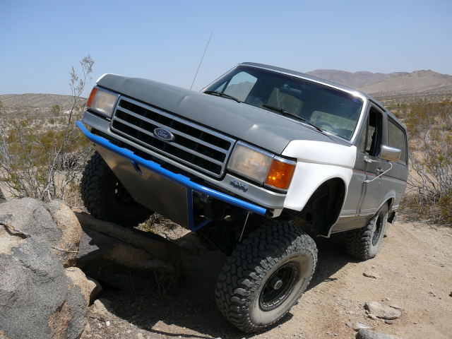 Ford Bronco with a blue bumper climbing over rocks and logs off-road