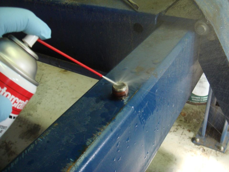 Close-up of a gloved hand spraying a bolt with lubricant from a red-tipped can