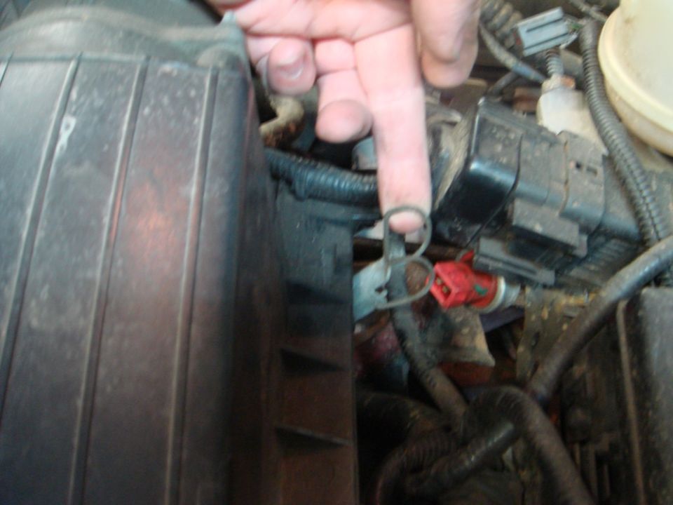 Close-up of engine bay with a finger pointing to a metal clip near a red electrical connector