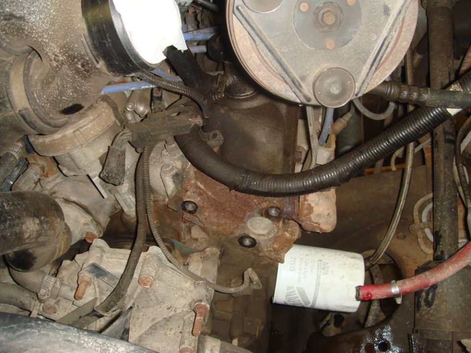Engine block with oil filter, hoses, and pulleys. Rusty metal and black hoses are visible