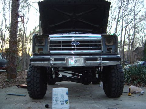 1986 Ford Bronco - This lifted Ford truck features a prominent chrome grille and oversized off-road tires, with its hood open for maintenance.