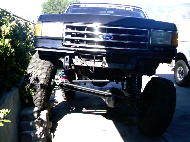 Black Ford Bronco with lifted suspension and large tires, front view. Another white truck is visible to the right