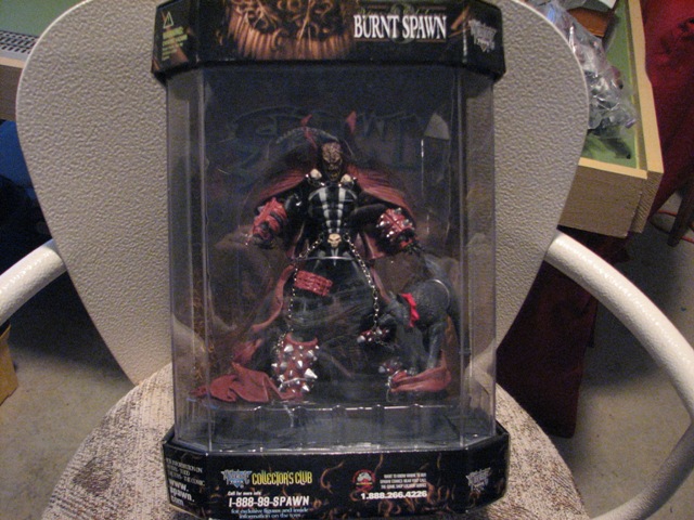 Action figure in clear plastic packaging with "BURNT SPAWN" text on top, placed on a chair