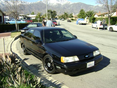 1991 Ford Taurus - A black sedan is parked on the side of a street with other vehicles and houses visible.