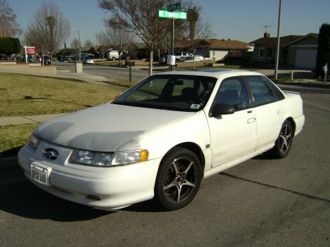 1991 Ford Taurus - A white 1991 Ford Taurus sedan with aftermarket wheels is parked on a residential street.
