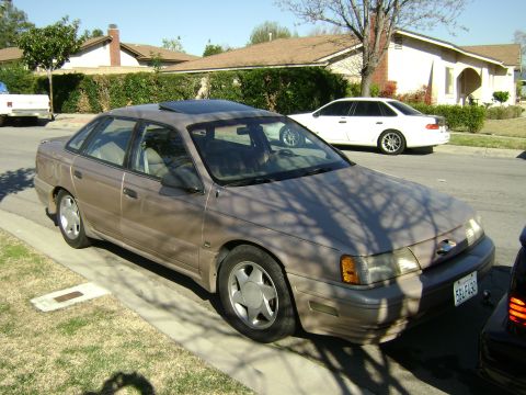 1991 Ford Taurus - A tan 1991 Ford Taurus is parked on the side of the road with houses and trees in the background.