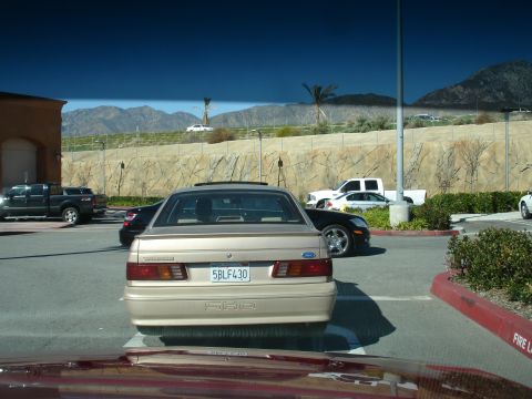 1991 Ford Taurus - A tan 1991 Ford Taurus is parked in a lot with other vehicles and mountains in the background.