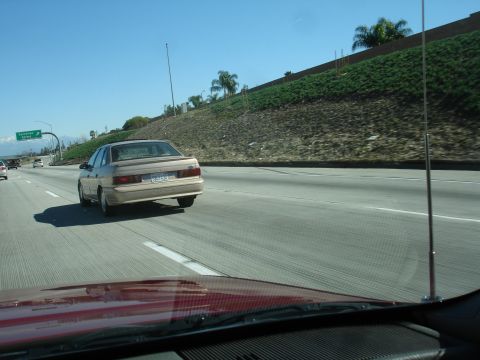 1991 Ford Taurus - A tan sedan travels down a sunny highway, its rear end visible from a following vehicle.