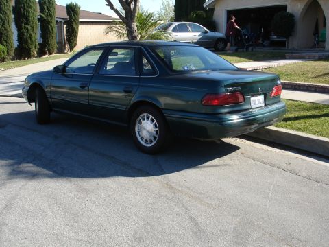 1991 Ford Taurus - The rear of a dark green sedan is visible, parked on a residential street with a house and another car in the background.