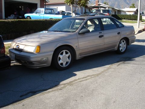 1991 Ford Taurus - A tan sedan with a sunroof is parked on a residential street next to a blue pickup truck.