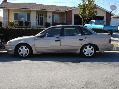 1991 Ford Taurus - A 1991 Ford Taurus sedan is parked in front of a house, with a blue pickup truck visible in the driveway.