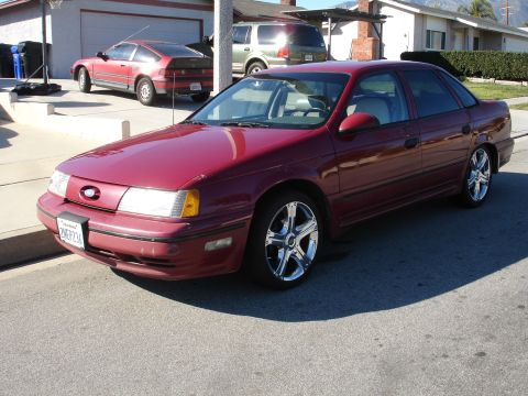 1991 Ford Taurus - A maroon 1991 Ford Taurus is parked in front of a suburban house, with other vehicles visible in the driveway.
