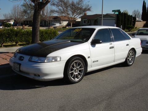 1991 Ford Taurus - A white sedan, possibly a 1991 Ford Taurus, is parked curbside with a contrasting black hood and aftermarket wheels.