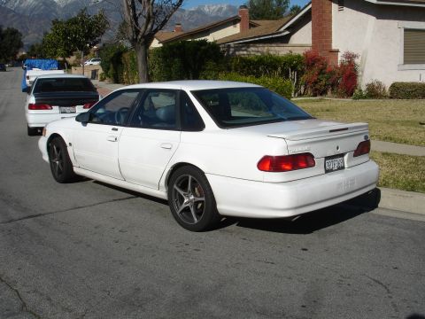 1991 Ford Taurus - A white sedan is parked on a residential street, with another vehicle behind it.