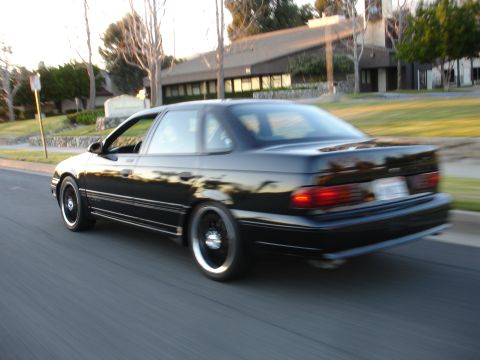 1991 Ford Taurus - A black 1991 Ford Taurus coupe is captured in motion on a suburban street during golden hour.