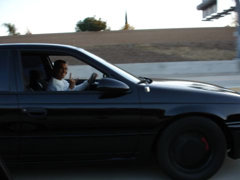 1991 Ford Taurus - Driver of a black sedan smiles and gives a peace sign while on the road.