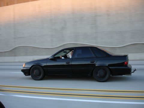 1991 Ford Taurus - A black 1991 Ford Taurus sedan is captured in profile while driving on a highway.