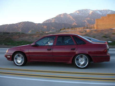 1991 Ford Taurus - A red sedan drives along a highway with mountains visible in the distance.