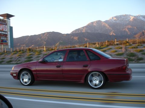 1991 Ford Taurus - A red 1991 Ford Taurus sedan is captured in a side profile shot while driving on a highway, with mountains in the background.