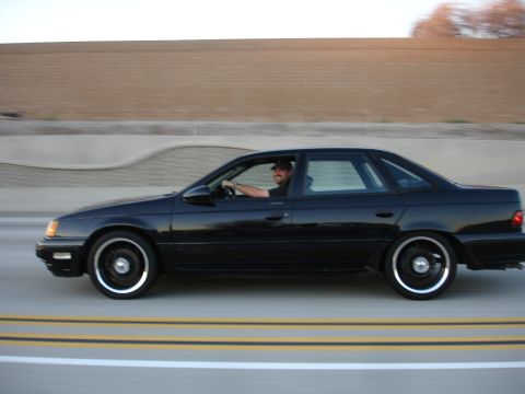 1991 Ford Taurus - A man drives a black sedan down a highway, with a blurred background indicating motion.
