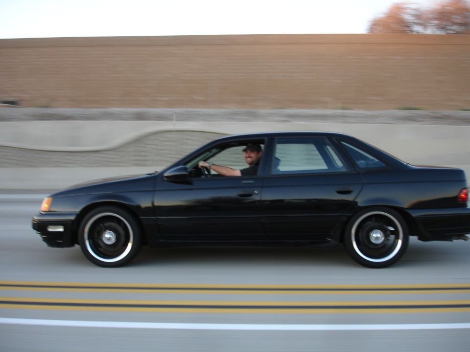 Side view of a black 1991 Ford Taurus sedan driving on a highway with a man behind the wheel