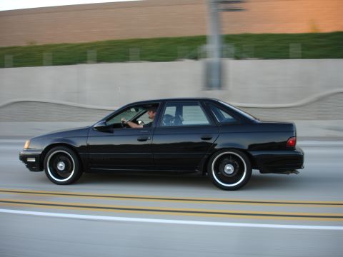 1991 Ford Taurus - A black 1991 Ford Taurus sedan is captured in motion on a highway, viewed from the side.