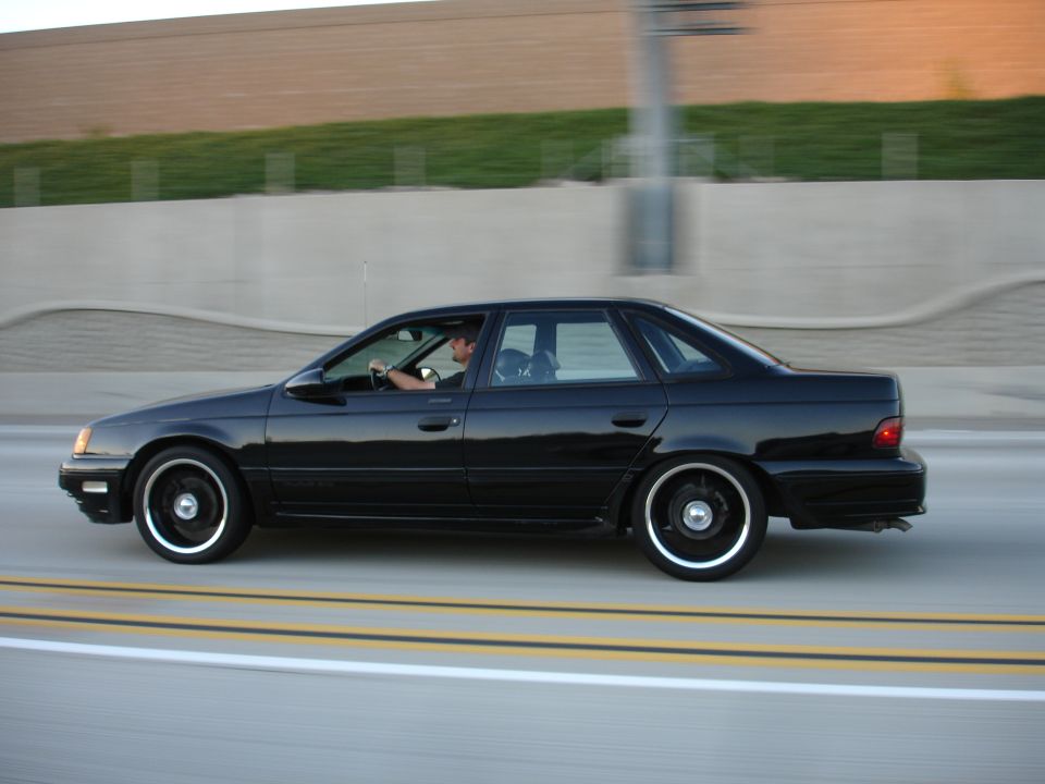 Black 1991 Ford Taurus sedan driving on a highway, side view