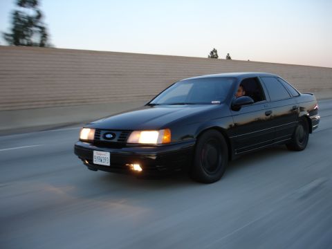 1991 Ford Taurus - A black 1991 Ford Taurus sedan is captured in motion on a highway during the golden hour.