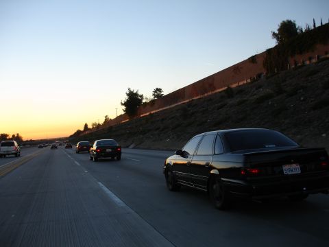 1991 Ford Taurus - A black sedan with a spoiler drives down the highway as the sun sets, casting a warm glow on the sky.