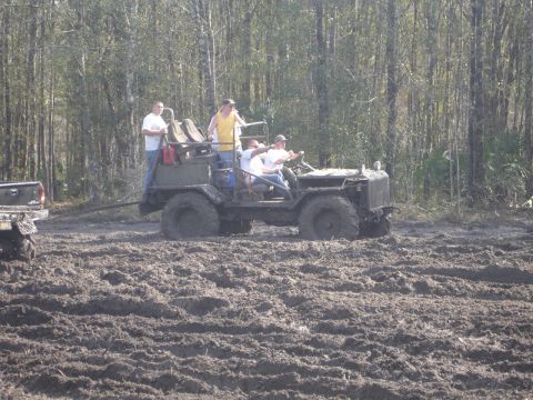 1989 Ford Bronco - An olive green off-road vehicle with several people aboard navigates a muddy terrain, with a pickup truck visible to the left.