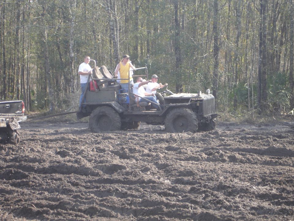 Olive green off-road vehicle with people in a muddy field, being towed by a pickup truck