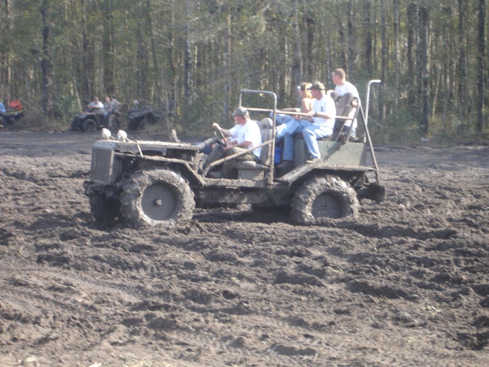 Mud bogging vehicle with passengers driving through thick mud, other ATVs visible in background