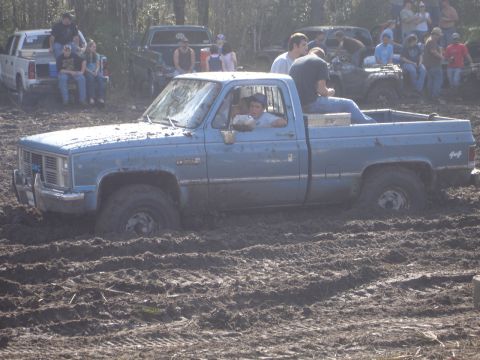 1989 Ford Bronco - A blue GMC pickup truck is stuck deep in the mud, with several people observing the scene.