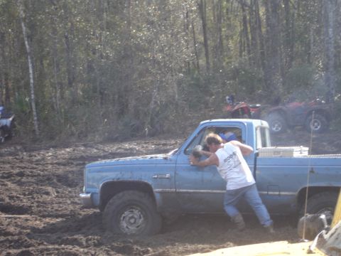 1989 Ford Bronco - A person attempts to free a blue pickup truck from thick mud while ATVs are visible in the wooded background.