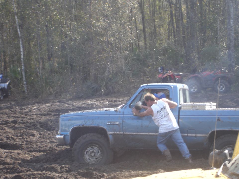 Blue pickup truck stuck in mud, driver pushing door. Other ATVs visible in background